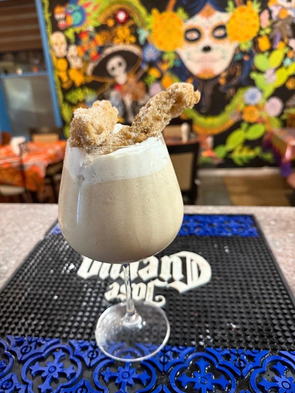 A creamy drink topped with churros in front of Hacienda’s Mexican artwork