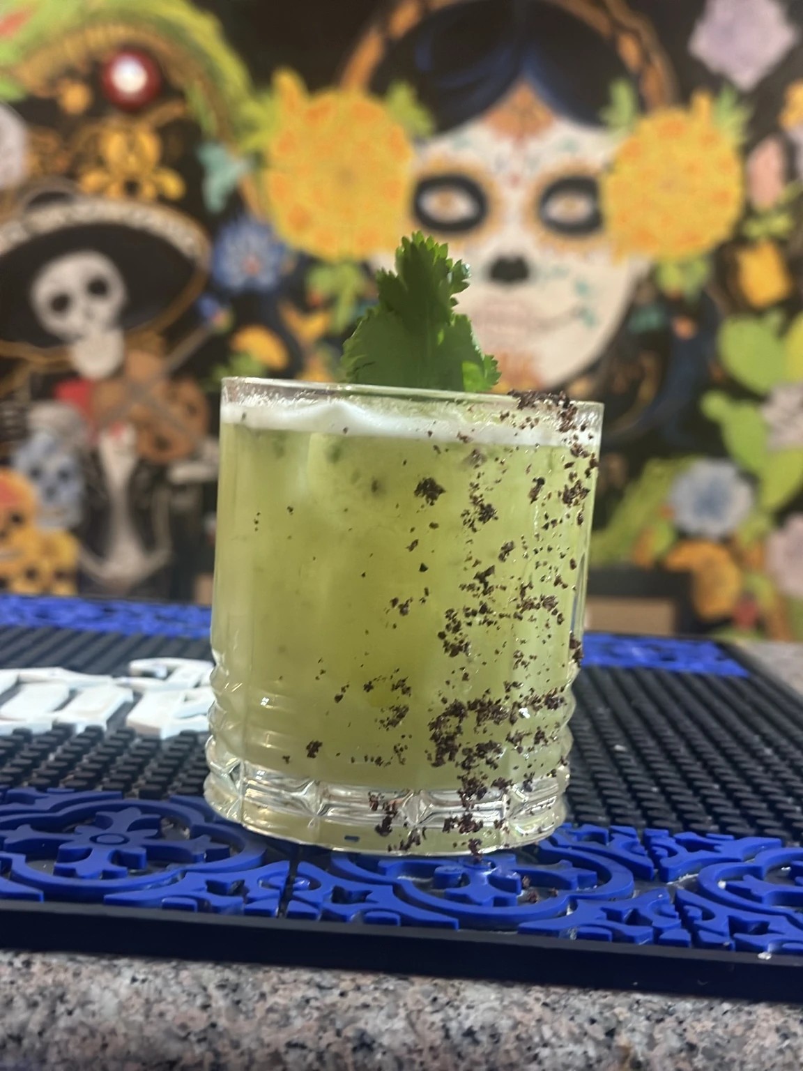 A green cocktail with a herb garnish and seasoned rim in front of Hacienda Santa Teresa’s Mexican artwork
