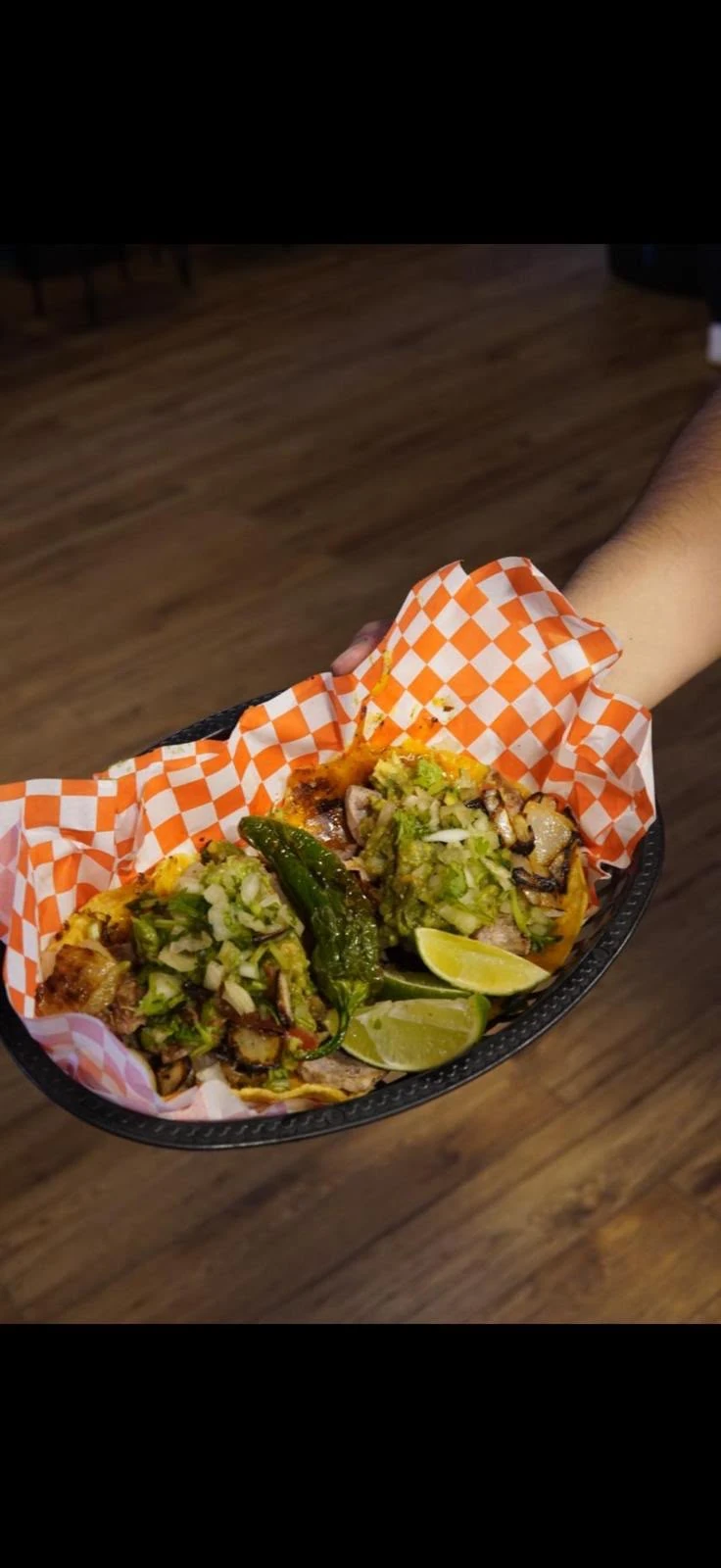 A basket of tacos topped with onion, cilantro, and guacamole, served with lime and a green pepper