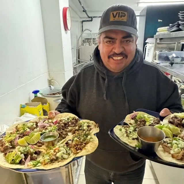 A smiling Hacienda team member holding two platters of tacos in the restaurant kitchen